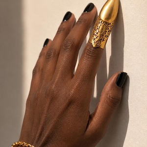 May include: A gold-toned metal finger armor ring featuring an intricate filigree cutout pattern on the base and a smooth, pointed cap covering the fingertip. The piece is worn alongside a simple gold-toned chain link bracelet on the wrist.