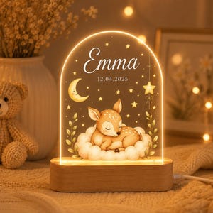Può includere: Una luce notturna ad arco in acrilico con base in legno, caratterizzata da un'illustrazione luminosa di un cerbiatto che dorme su una nuvola. Il design include una mezzaluna, stelle e il testo Emma 12.04.2025. La luce emette un bagliore caldo.
