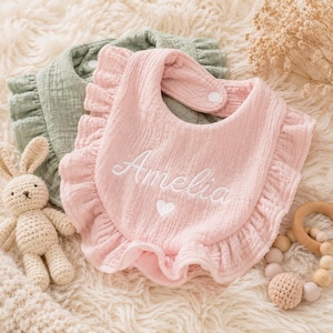 May include: A light pink muslin bib with ruffled edges and the embroidered name Amelia above a small white heart. A second sage green muslin bib is layered underneath. Both bibs feature a snap closure at the neck for secure fastening.
