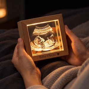 Può includere: Una scatola luminosa quadrata in legno che mostra un'ecografia fetale illuminata. La cornice è realizzata in legno chiaro e presenta la scritta Baby Olivia e un piccolo simbolo a forma di cuore incisi sul bordo inferiore della cornice.