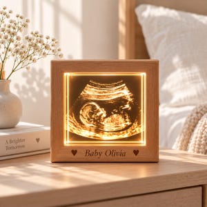 May include: A square wooden light box featuring an illuminated ultrasound scan of a fetus. The frame is made of light-toned wood and includes the engraved text Baby Olivia flanked by two small heart symbols at the bottom centre of the frame.