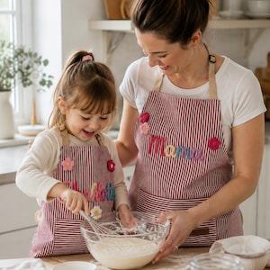 Può includere: Due grembiuli abbinati con un motivo a righe sottili rosse e bianche e lacci beige al collo. Il grembiule più grande mostra la scritta ricamata Mama in lettere colorate, mentre quello più piccolo riporta il nome ricamato Charlotte.