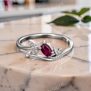 Könnte beinhalten: Ein silberfarbener Ring mit einem zentralen, oval geschliffenen, tiefroten Rubin. Die Ringschiene ist mit kleinen, klaren Steinen in Blattform sowie einer geschwungenen Reihe von Pavé-Steinen an einer Seite verziert.