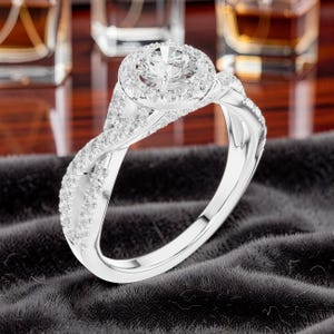 May include: A silver-toned ring featuring a large round-cut center stone surrounded by a halo of smaller pavé-set stones. The band has a twisted design, with both strands encrusted with numerous small, clear, sparkling stones along the shank.