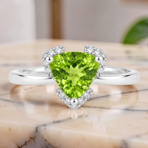 Peut inclure: Une bague en argent avec une grande pierre péridot vert lime taillée en triangle au centre, entourée d'un halo de petites pierres blanches rondes et transparentes serties le long des bords de la monture triangulaire sur un anneau poli.