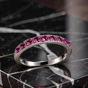 Könnte beinhalten: Ein silberfarbener Ring mit einer Reihe kleiner, rund geschliffener, leuchtend magentafarbener Edelsteine, die entlang der oberen Hälfte des Rings eingefasst sind. Der Ring hat eine polierte metallische Oberfläche und eine glatte Innenseite.