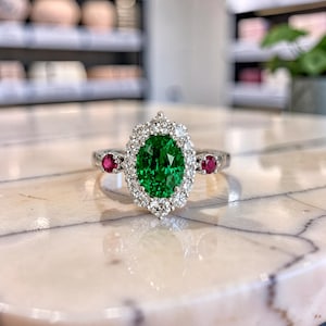 Könnte beinhalten: Ein silberner Ring mit einem zentralen, oval geschliffenen grünen Edelstein, der von einem Kranz aus kleinen runden weißen Diamanten umgeben ist. Die Ringschiene enthält auf jeder Seite des zentralen Steins einen runden roten Edelstein.