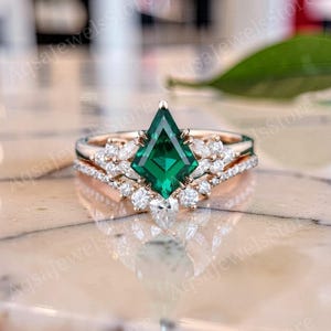 Könnte beinhalten: Ein Ring-Set aus Roségold mit einem zentralen, drachenförmigen grünen Edelstein, der von weißen Steinen im Marquise- und Rundschliff flankiert wird. Ein passender Ring mit kleinen runden weißen Steinen ergänzt das Design zu einer spitzen Form.