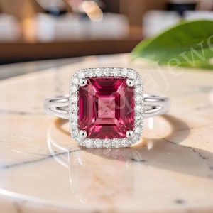 Könnte beinhalten: Ein silberner Ring mit einem großen, rechteckigen, dunkelrosa Edelstein in der Mitte, umgeben von einem Kranz aus kleinen, klaren, rund geschliffenen Steinen. Die Ringschiene ist aus poliertem Silber und hat ein geteiltes Design am Ansatz.