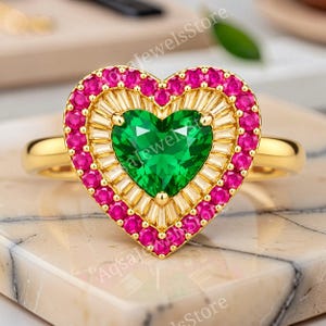 Könnte beinhalten: Ein Goldring mit einem herzförmigen grünen Mittelstein, umgeben von einem Heiligenschein aus klaren Baguette-Steinen und einem äußeren Rand aus runden rosa Steinen. Das Band ist aus glattem Gold und die Fassung ist herzförmig gestaltet.