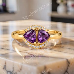 Könnte beinhalten: Ein Goldring mit zwei violetten Amethysten im Trillantschliff, die nebeneinander angeordnet sind und von einem Kranz aus kleinen weißen Diamanten im Rundschliff umgeben werden. Der Ring ist aus poliertem Gold und trägt den Text AqsaJewelsStore.