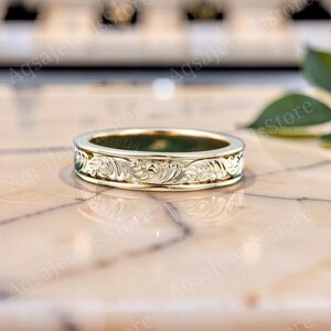 Könnte beinhalten: Ein goldener Bandring mit einem komplizierten, durchgehenden gravierten Rankenmuster auf der Außenfläche. Das Metall hat eine polierte, reflektierende Oberfläche, die das Licht einfängt und die detaillierten, wirbelnden blumenartigen Schnitzereien hervorhebt.