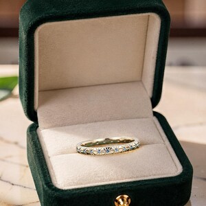 Könnte beinhalten: Ein goldener Bandring mit einer Reihe eingravierter Sternenmuster, die jeweils mit einem kleinen blauen Edelstein besetzt sind. Der Ring befindet sich in einer offenen, dunkelgrünen Schmuckschatulle aus Samt mit cremefarbenem Innenfutter.