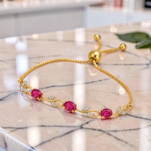 Könnte beinhalten: Ein verstellbares Armband in Goldoptik mit drei oval geschliffenen, leuchtend pinkfarbenen Steinen in einer zarten, rankenförmigen Metallfassung. Das Design enthält kleine, klare Akzentsteine, die in die goldenen Blätter eingelassen sind.