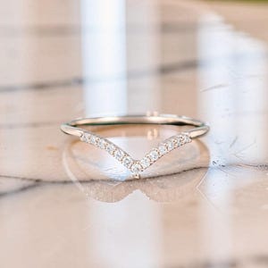 Könnte beinhalten: Ein V-förmiger Ring aus poliertem, silberfarbenem Metall mit einer Reihe kleiner, klarer, rund geschliffener Steine, die entlang des oberen Chevron-Abschnitts des Rings eingelassen sind und ein zartes, symmetrisches Design mit reflektierender Oberfläche bilden.