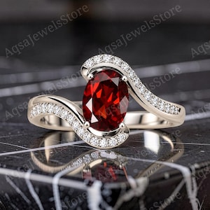 May include: A silver-toned ring featuring a central oval-cut deep red garnet gemstone. The band has a bypass design with two curving arms, each pavé-set with small clear round stones that extend from the centre setting along the shank.