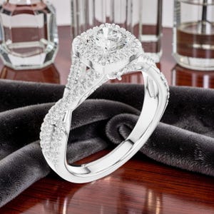 May include: A silver-toned ring featuring a large round centre stone surrounded by a square halo of smaller stones. The band has a twisted design encrusted with numerous tiny clear stones that extend along the sides of the ring setting.