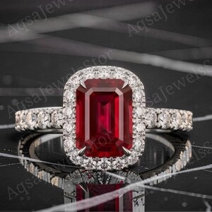 Può includere: Un anello color argento con una grande pietra preziosa rossa rettangolare a taglio smeraldo al centro. La pietra è circondata da un alone di piccole pietre rotonde trasparenti, con ulteriori pietre incastonate lungo la fascia su entrambi i lati.