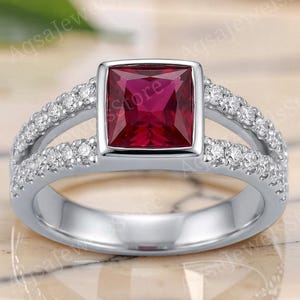 Könnte beinhalten: Ein silberfarbener Ring mit einem quadratisch geschliffenen, tiefroten Edelstein in der Mitte. Die Ringschiene ist zweigeteilt, wobei jede Reihe mit kleinen, klaren, rund geschliffenen Steinen besetzt ist, die sich zu den Seiten hin erstrecken.