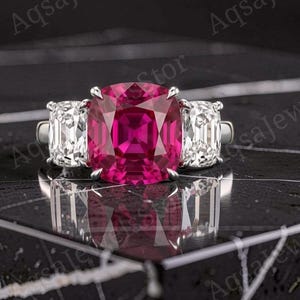 May include: A three-stone ring featuring a large, cushion-cut, vibrant pink centre gemstone flanked by two smaller, rectangular, clear white side stones. The stones are set in a polished silver-toned metal band with four prongs for the centre stone.