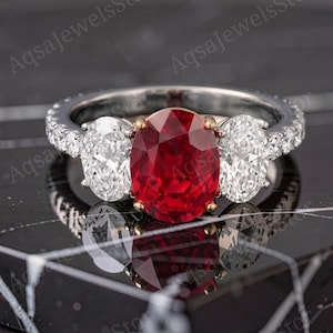 Könnte beinhalten: Ein Ring mit drei Steinen, bestehend aus einem großen oval geschliffenen roten Edelstein in der Mitte, flankiert von zwei kleineren oval geschliffenen klaren weißen Steinen. Die Ringschiene ist aus silberfarbenem Metall und seitlich besetzt.