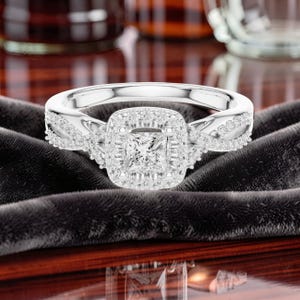 May include: A silver-toned ring featuring a central princess-cut stone surrounded by a halo of smaller round stones. The band has a twisted design encrusted with tiny round stones on both sides, creating a sparkling and detailed appearance.