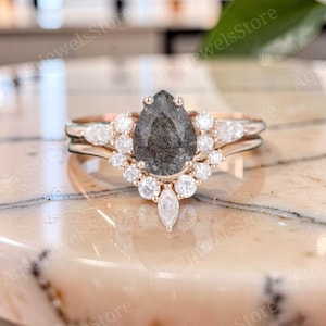 Könnte beinhalten: Ein Ring aus Roségold mit einem zentralen birnenförmigen Rutilquarz mit dunklen Einschlüssen. Der Stein ist von einem Kranz aus kleinen runden und marquiseförmigen weißen Steinen in V-Form umgeben, gefasst auf einer polierten Roségoldschiene.
