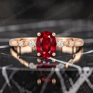 Könnte beinhalten: Ein Ring aus Roségold mit einem zentralen, oval geschliffenen, tiefroten Rubin, der von vier Krappen gehalten wird. Die Ringschiene zeigt auf jeder Seite zwei kleine, rund geschliffene weiße Diamanten in einem gedrehten Metalldesign.