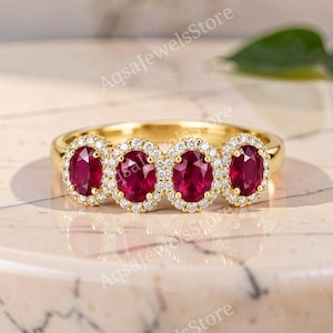 Könnte beinhalten: Ein Goldring mit vier oval geschliffenen, tiefroten Rubinen in einer Reihe. Jeder Rubin ist von einem Heiligenschein aus kleinen, klaren, rund geschliffenen weißen Steinen umgeben. Der Text AqsaJewelsStore ist im Hintergrund zu sehen.