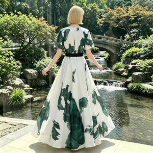 May include: A floor-length dress featuring a white base with dark green abstract brushstroke patterns, short sleeves, and a solid black waistband. The garment has a full, flowing skirt that reaches the ground, creating a wide, flared silhouette.