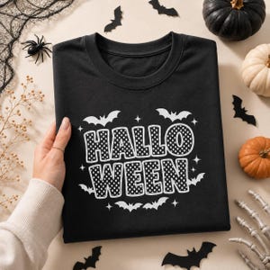 May include: A black t-shirt featuring the word HALLOWEEN in large, white, polka-dotted block letters. The design is surrounded by small white bat silhouettes and four-pointed star shapes, printed on the center of the fabric.