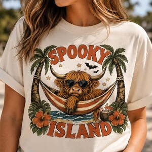 May include: A cream-colored t-shirt featuring a graphic of a brown Highland cow wearing black sunglasses while lounging in a hammock between two palm trees. The text reads SPOOKY ISLAND, accompanied by small stars, bats, and hibiscus flowers.
