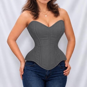 May include: A charcoal gray strapless corset top featuring a sweetheart neckline and vertical seam detailing throughout the bodice. The garment is fitted with a structured design and a pointed hemline, paired with dark blue denim jeans.