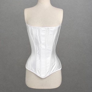 May include: A strapless white corset featuring a smooth, satin-like finish with vertical boning channels throughout the bodice. The garment has a straight neckline and a pointed hemline at the center front, displayed on a mannequin torso.