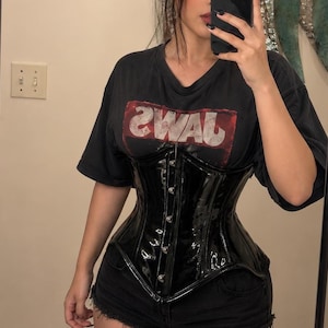 Puede incluir: Una camiseta negra de manga corta con un gráfico rojo y blanco con el texto JAWS, usada con un corsé de vinilo negro brillante que tiene un cierre frontal con broches metálicos plateados y un dobladillo en punta en el centro inferior.