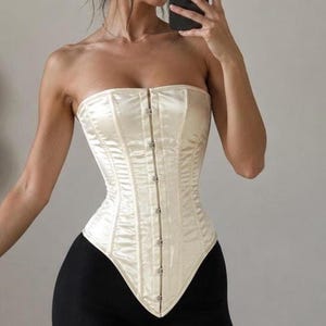 May include: A strapless cream-colored satin corset featuring a vertical row of silver-toned metal hook-and-eye closures down the center front. The garment has a pointed hemline that extends downward at the front and is constructed with vertical seams.