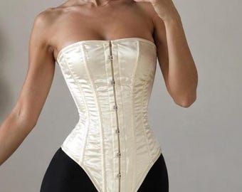 May include: A strapless cream-colored satin corset featuring a vertical row of silver-toned metal hook-and-eye closures down the center front. The garment has a pointed hemline that extends downward at the front and is constructed with vertical seams.