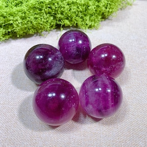 May include: Five polished, spherical purple fluorite crystal spheres. The spheres are arranged in a circular pattern on a neutral-toned surface, with a vibrant green moss-like element in the background. The spheres have a glossy finish.