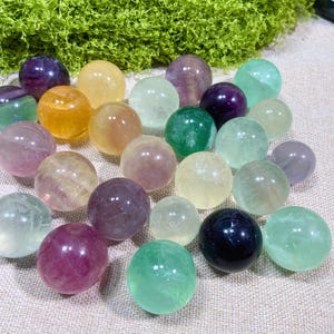 May include: A collection of polished, spherical fluorite stones in various translucent colours including shades of purple, green, yellow, and clear. The smooth, round mineral spheres are arranged in a cluster, showcasing natural colour variations.