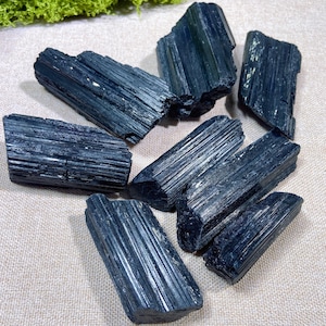May include: A collection of seven raw black tourmaline crystals, each featuring a distinct elongated shape with vertical striations and a dark, opaque, glossy surface. These natural mineral specimens are arranged in a scattered formation.
