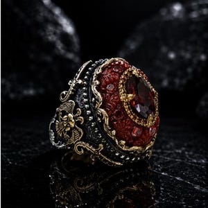 Puede incluir: Un anillo en tonos plata y oro con una piedra preciosa de color rojo oscuro de corte ovalado en el centro, rodeada de piedras rojas más pequeñas. La banda presenta intrincados detalles metálicos, incluyendo un águila y motivos florales.
