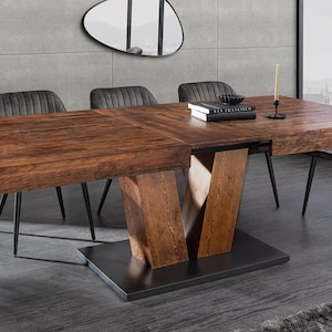 Puede incluir: Una mesa de comedor rectangular de madera con un acabado marrón oscuro. La mesa tiene una base única en forma de V y está rodeada de sillas grises tapizadas con patas de metal negro. Un libro y un candelabro están sobre la mesa.