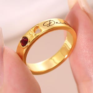 Puede incluir: Un anillo de banda en tono dorado que presenta una pequeña piedra preciosa redonda de color rojo oscuro y una forma de corazón recortada. La superficie exterior del anillo tiene grabado el texto "Daisy" en cursiva negra, junto al corazón.