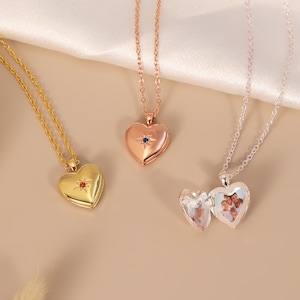 May include: Three heart-shaped locket necklaces in gold, rose gold, and silver tones. The gold and rose gold lockets feature a small starburst design with a central gemstone. The silver locket is open, revealing a small photograph of a family inside.