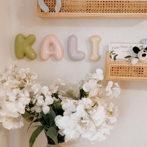 May include: Wall-mounted balloon-style letters spelling KALI in olive green, soft pink, lavender, and cream. Above are two wooden shelves with cane webbing holding plush toys and books. Below is a grey vase with white hydrangea flowers.