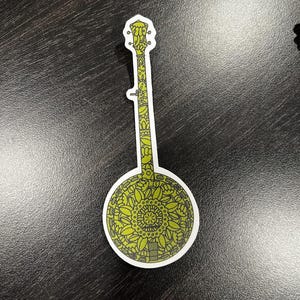 Puede incluir: Una pegatina de banjo verde y negro con un diseño floral detallado. El banjo tiene un cuerpo redondo y un cuello largo.