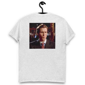 Peut inclure: T-shirt gris clair avec une image carrée d'un homme en costume et cravate. L'homme regarde le spectateur avec un léger sourire. Le t-shirt est suspendu à un cintre noir.