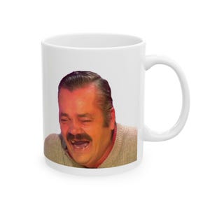 Peut inclure: Mug en céramique blanche avec un mème d'un homme riant avec une moustache. L'homme porte un pull beige. La tasse a une anse blanche et est posée sur un fond blanc.