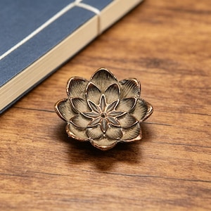 May include: A small, metallic lotus flower ornament with a weathered bronze finish. The object features multiple layers of sculpted, pointed petals radiating from a central point, creating a dimensional, floral shape with dark, recessed detailing.