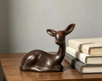 May include: A dark brown, smooth-surfaced figurine of a resting deer with its head turned slightly to the side. The deer is depicted in a seated position with its legs tucked underneath its body, featuring pointed ears and a slender neck.